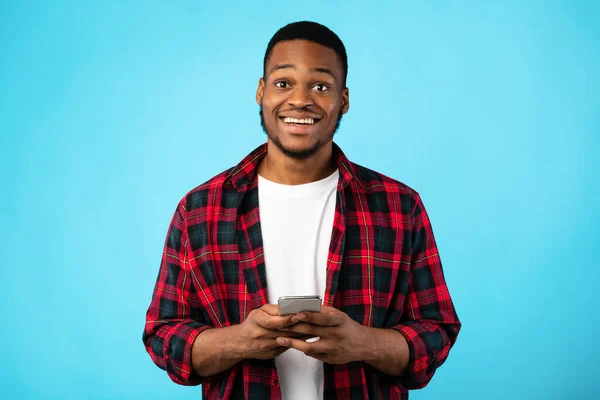 Chico negro alegre usando Smartphone sonriendo de pie sobre fondo azul — Foto de Stock