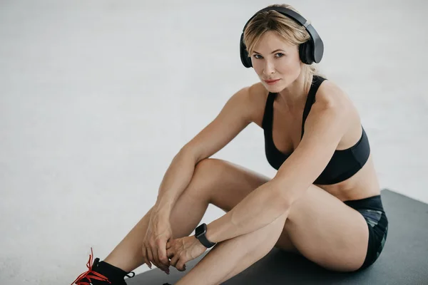 Mujer adulta en ropa deportiva y grandes auriculares modernos se sienta en el suelo en casa durante la cuarentena —  Fotos de Stock