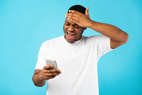 Homem negro desesperado segurando telefone leitura más notícias, fundo azul — Fotografia de Stock