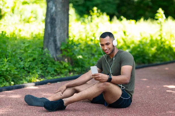 Zrelaksowany czarny mężczyzna siedzi na torze do joggingu i słucha muzyki ze smartfona w parku — Zdjęcie stockowe