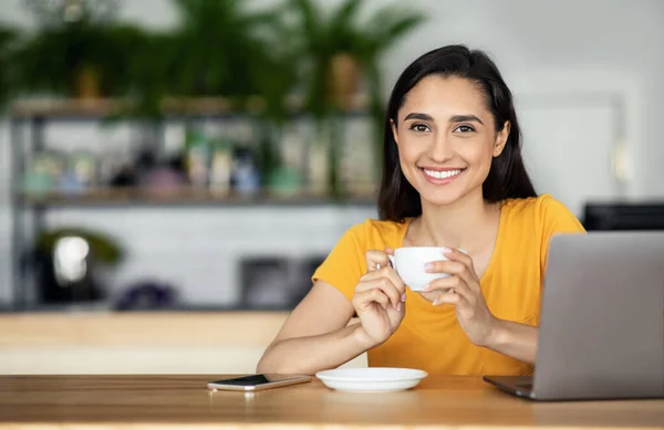 Usmívající se hezká dívka pije čaj v kavárně, pomocí notebooku — Stock fotografie