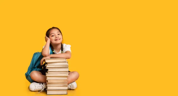 Azjatycki elementarny student dziewczyna siedzi przy książki stosu, żółte tło — Zdjęcie stockowe