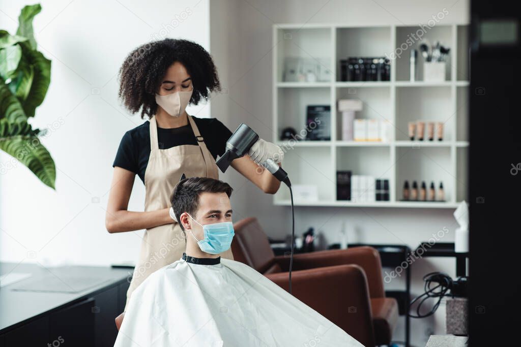 Black hairdresser making new hairstyle for client, both in protective masks