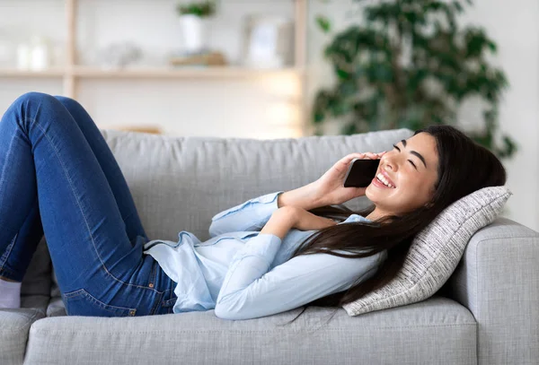 Позитивная азиатка расслабляется на диване, разговаривает по телефону с друзьями — стоковое фото