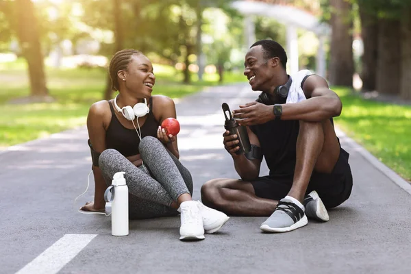 Alegre pareja africana descansando después de hacer ejercicio al aire libre, sentada en el camino en el parque —  Fotos de Stock