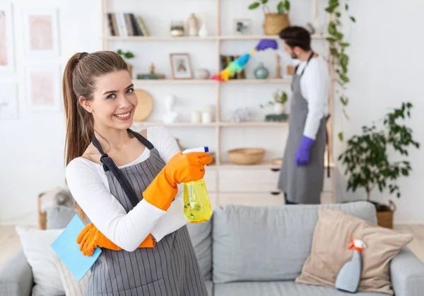 A limpar a casa juntos. Mulher sorridente com spray de perto, aponta para marido ocupado — Fotografia de Stock