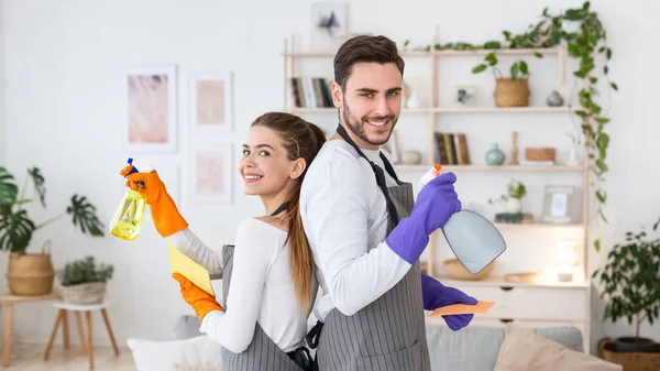 Gran limpieza en casa. Marido y esposa sonrientes en delantales y guantes de goma con aerosoles en la sala de estar —  Fotos de Stock