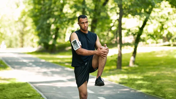 Yakışıklı siyah sporcu yaz parkında koşudan sonra bacağını esnetiyor.