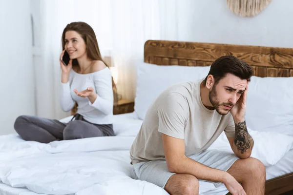 Problemas familiares. Mulher falando ao telefone, marido descontente senta-se na cama e segura a cabeça com a mão — Fotografia de Stock