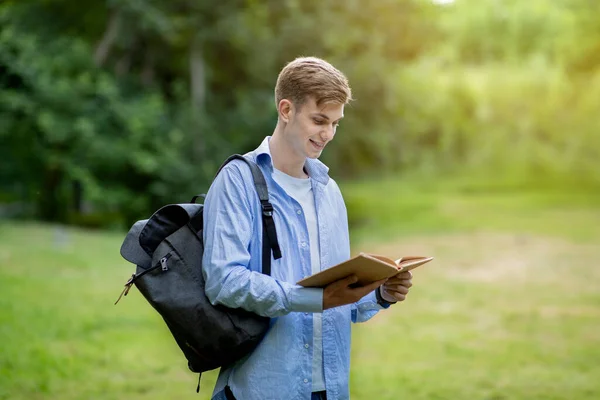 Концепция обучения. Красавчик-студент читает книгу на открытом воздухе, отдыхает в кампусе — стоковое фото
