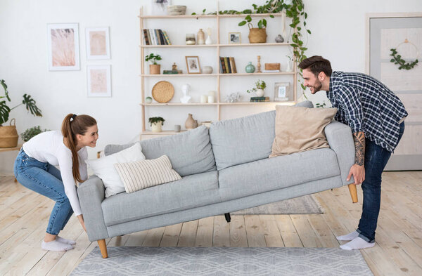 Moving or permutation furniture at home. Happy guy and girl raise sofa in interior of living room