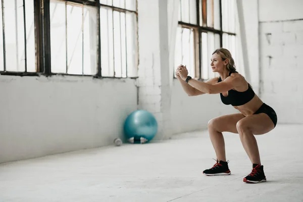 Sportovní štíhlá žena ve sportovním oblečení s fitness tracker cvičení squat cvičení ve studiu za denního světla — Stock fotografie