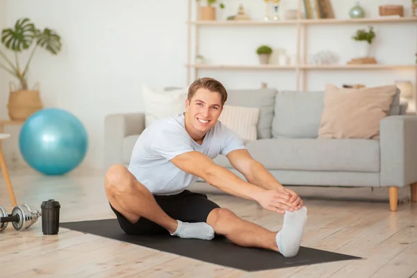 Spor giyim sektöründe mutlu bir adam oturma odasının iç kısmında halterler ve spor fincanlarıyla paspasın üzerinde geriniyor. — Stok fotoğraf