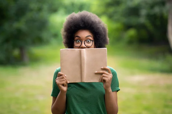 Книжковий хробак. смішна афроамериканська студентка стоїть на відкритому повітрі обличчя з книгою — стокове фото