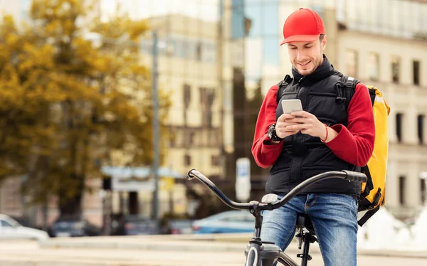 Kurier za pomocą aplikacji na smartfonie siedzi na rowerze w mieście — Zdjęcie stockowe