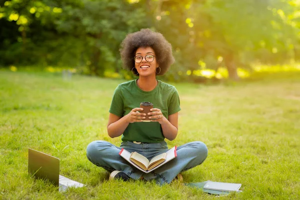 Чорна студентка відпочиває в кампусі коледжу, сидить на галявині з кавою — стокове фото