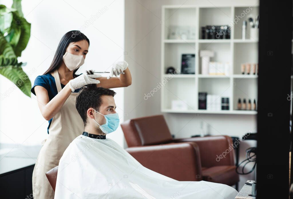 Young female master cutting hair for male client, both in protective masks.