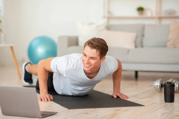 Online-Training zu Hause. Lächelnder Typ in Sportbekleidung macht Liegestütze und blickt im Wohnzimmer auf Laptop — Stockfoto