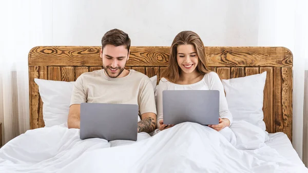 Szabadidő együtt. Fiatal férj és feleség ül az ágyon, és laptopokat néznek — Stock Fotó