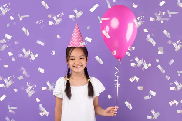 Asiatisches Mädchen hält Luftballon unter fallendem Konfetti, lila Hintergrund — Stockfoto