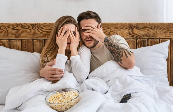 Marido assustado e esposa olhos cobertos com as mãos no interior do quarto com controle remoto e pipoca — Fotografia de Stock