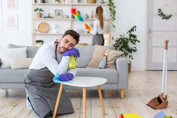 Tareas domésticas y cansancio. Hombre en delantal y guantes durmiendo en sus rodillas y mesa, esposa limpia muebles — Foto de Stock