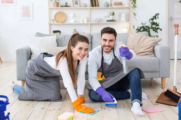 Fácil limpieza con placer. Feliz hombre y mujer lavan el piso con suministros de limpieza en la sala de estar —  Fotos de Stock