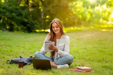 Neşeli genç öğrenci kız elinde not defteriyle çimlerde oturuyor ve not alıyor.