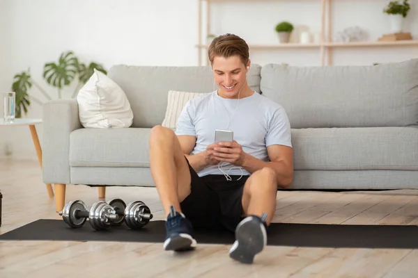 Atrakcyjny młodzieniec ze słuchawkami w odzieży sportowej siedzi na macie na podłodze z hantlami i patrzy na smartfona — Zdjęcie stockowe