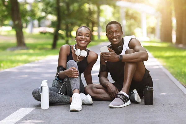 Deportiva pareja africana posando después de entrenar al aire libre, sentada en el camino en el parque —  Fotos de Stock