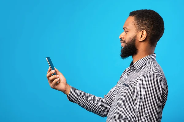 Afro man menggunakan ponsel pada dinding biru, tampilan samping — Stok Foto