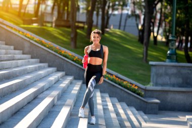 Neşeli sportif kız su içiyor, parkta merdivenlerde duruyor.