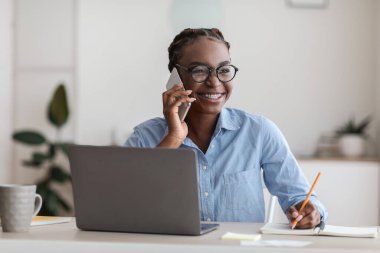 Siyah kadın ofis müdürü cep telefonuyla konuşuyor ve iş yerinde not alıyor.