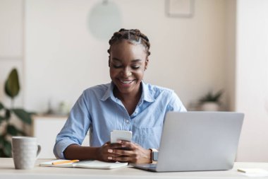 Ofiste akıllı telefon kullanan gülümseyen Afro-Amerikan iş kadını.