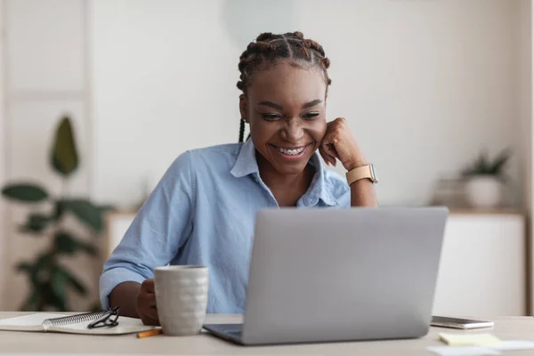 Sonriente empleada negra usando computadora portátil y beber café en el lugar de trabajo de la oficina — Foto de Stock