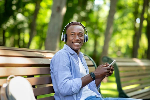 Тисячолітній чорний чоловік сидить на лавці в парку і слухає музику з мобільного телефону — стокове фото