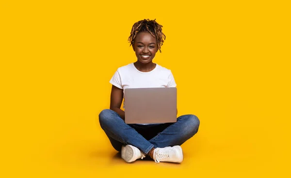 Black girl sitting on floor, using laptop, yellow background — Stock Photo, Image