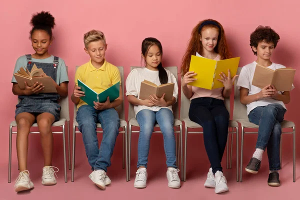 Sokszínű gyerekek tankönyvekkel és jegyzetfüzetekkel, székeken ülve, készülve az iskolai leckére rózsaszín háttérrel — Stock Fotó