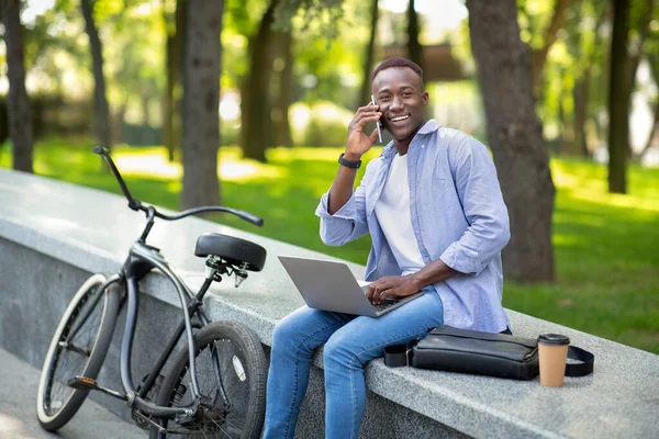 Homem negro feliz com bicicleta falando no smartphone enquanto usa seu laptop no parque no dia de verão, espaço de cópia — Fotografia de Stock