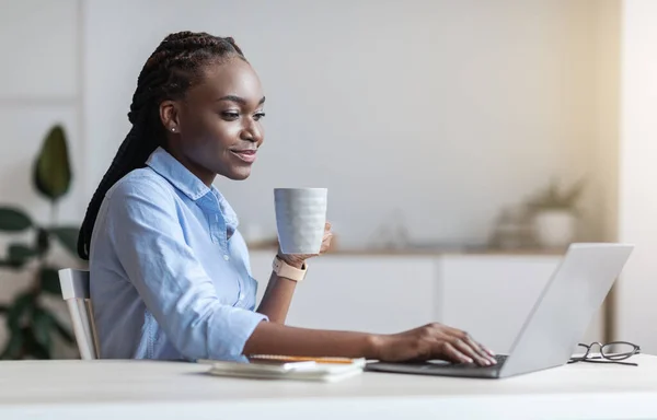 Ung svart affärskvinna dricker morgonkaffe medan du arbetar på laptop på kontoret — Stockfoto