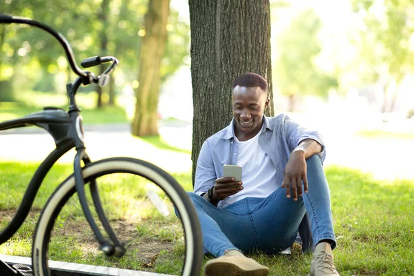 Millennial homem preto com bicicleta sentado perto da árvore e usando telefone celular no parque de verão — Fotografia de Stock