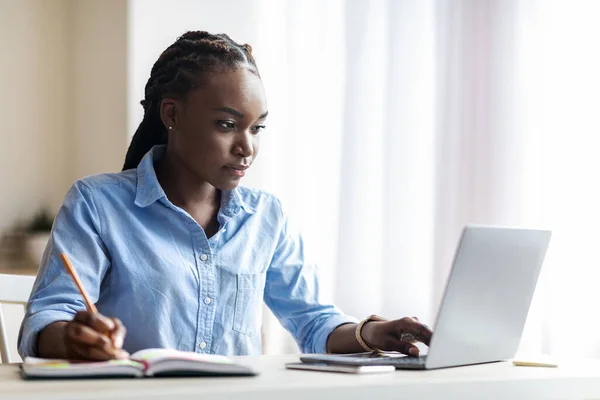 Entrepreneure africaine confiante travaillant sur ordinateur portable et prenant des notes au bureau — Photo