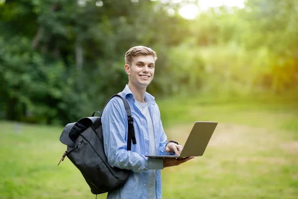 Happy student guy with laptop and backpack posing outdoors at college campus