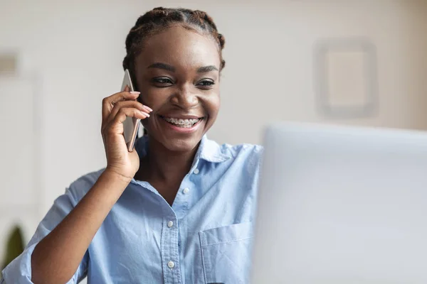 Comunicazione commerciale. Donna d'affari nera che parla sul cellulare e utilizza il computer portatile sul posto di lavoro — Foto Stock