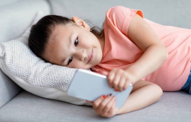 Küçük Koreli kız cep telefonu kullanıyor. Evdeki koltukta yatıyor.