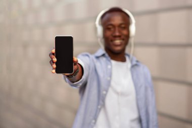 Milenyum siyahi kulaklıklı bir adam akıllı telefonu açık hava ekranlı, seçici odaklı gösteriyor. Tasarım için model