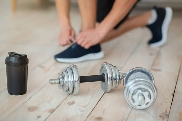 Training with dumbbells at home. Guy tying shoes, near cup and sport equipment — Stock Photo, Image