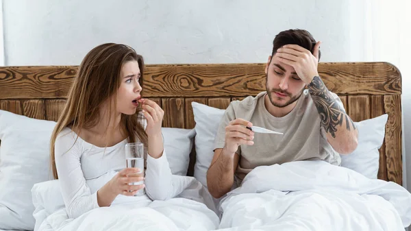 Vrouw die pil drinkt, een glas water vasthoudt en naar haar man kijkt, haar voorhoofd aanraakt en naar de thermometer kijkt — Stockfoto