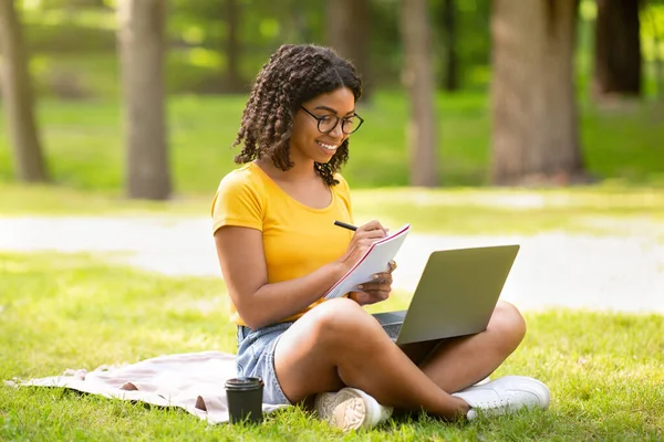 Zdalna edukacja. Radosny czarny lady robi notatki podczas oglądania webinarium na laptopie w parku — Zdjęcie stockowe