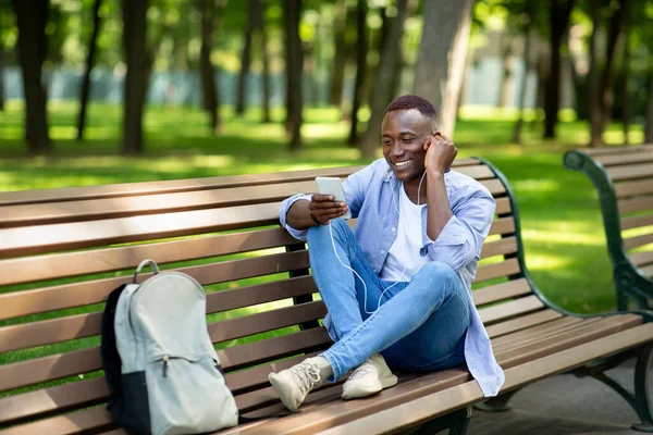 Веселий афроамериканець сидить на лавці і насолоджується музикою на мобільному телефоні в парку, вільне місце — стокове фото